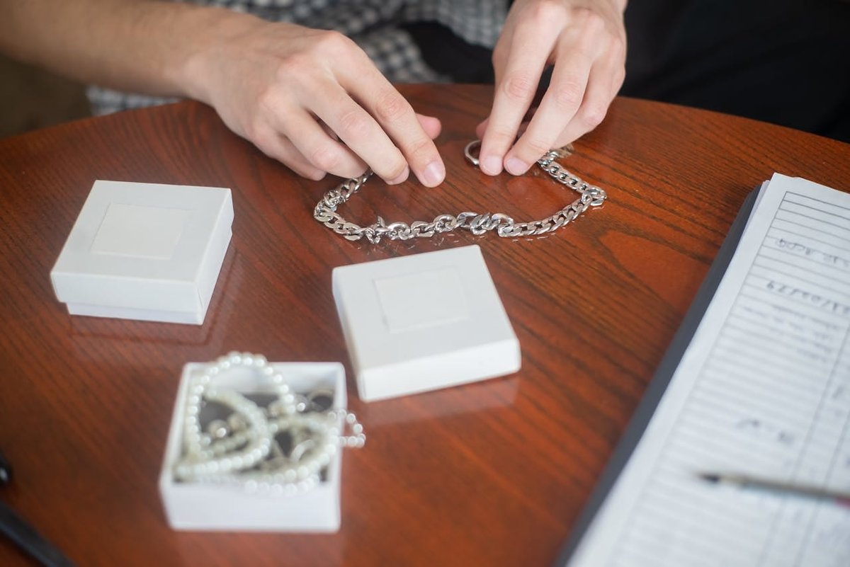 שור הזהב Hands arranging a silver chain necklace on a wooden table with jewelry boxes, indoors.